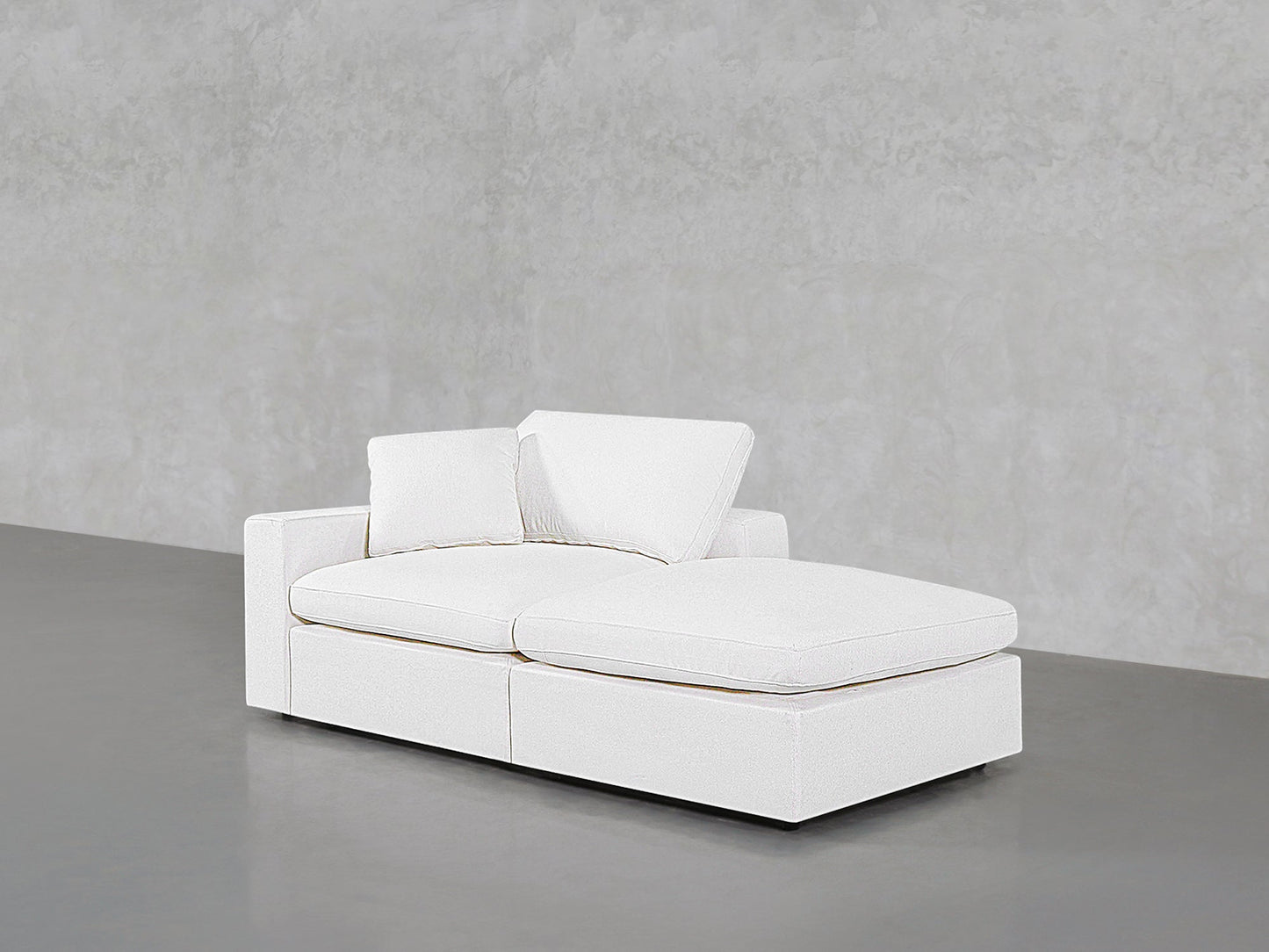A 2-Seat Modular Lounger Loveseat in modern white upholstery with two matching cushions is placed on a smooth grey floor against a textured light grey wall.