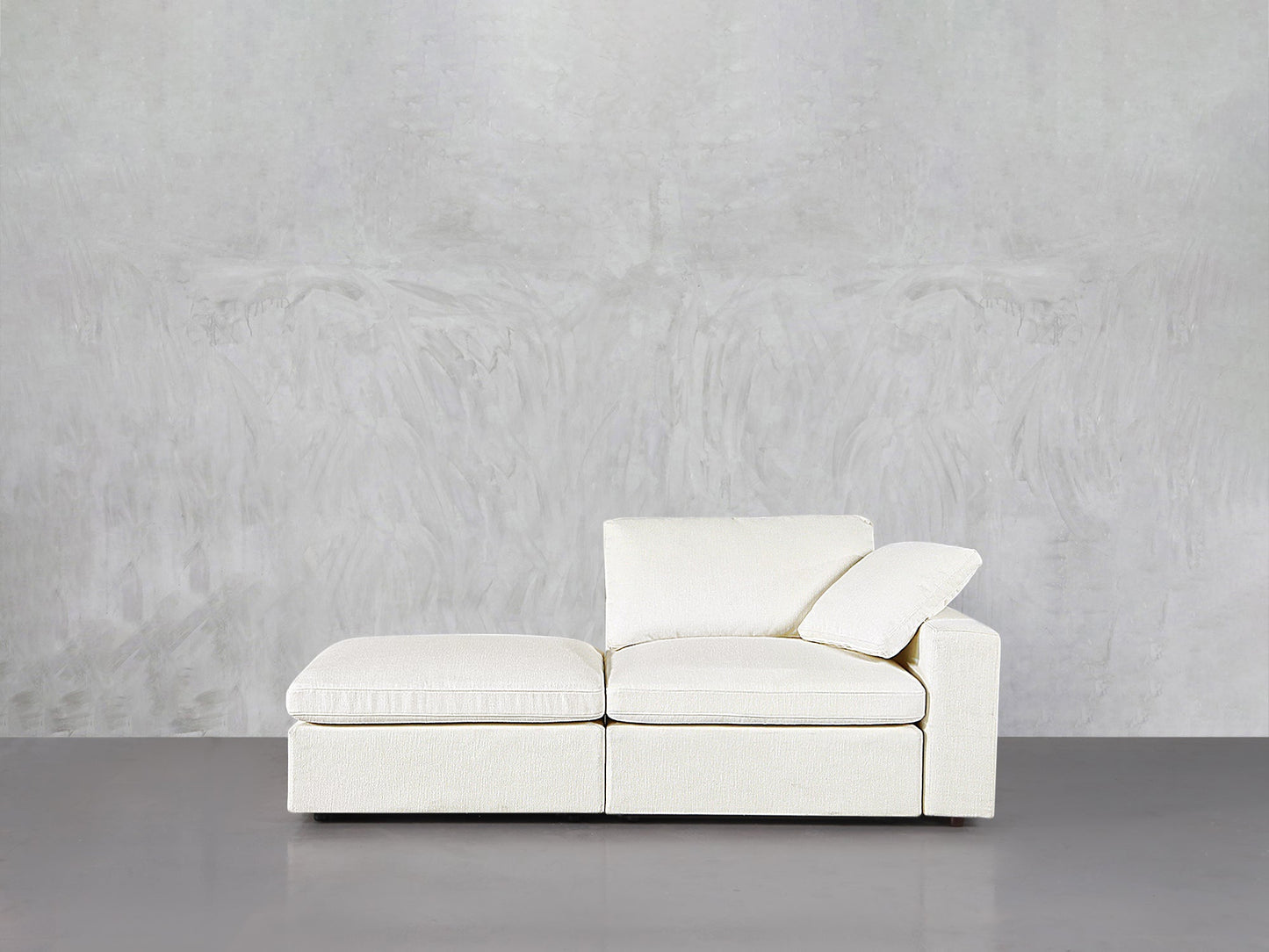 The 2-Seat Modular Lounger Loveseat in off-white, featuring a right armrest and loose cushion, sits against a light gray textured wall on a smooth gray floor.