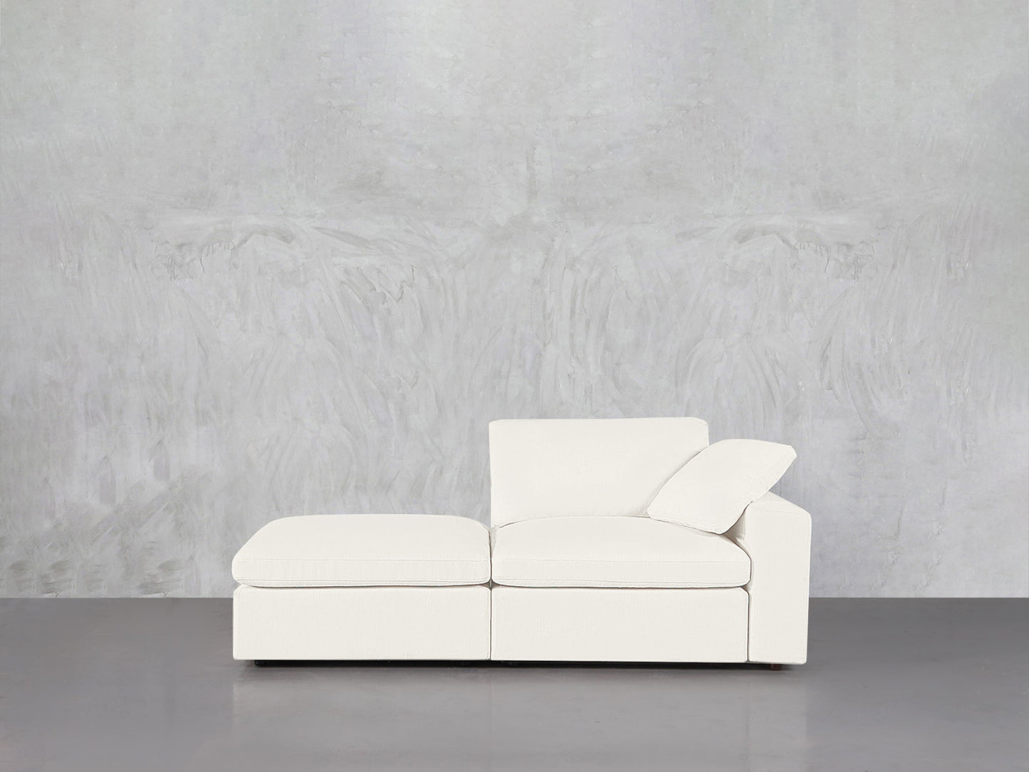A 2-Seat Modular Lounger Loveseat in white, paired with an ottoman, sits against a light gray textured wall on a smooth gray floor. One cushion is angled for support in this minimalistic, uncluttered space.
