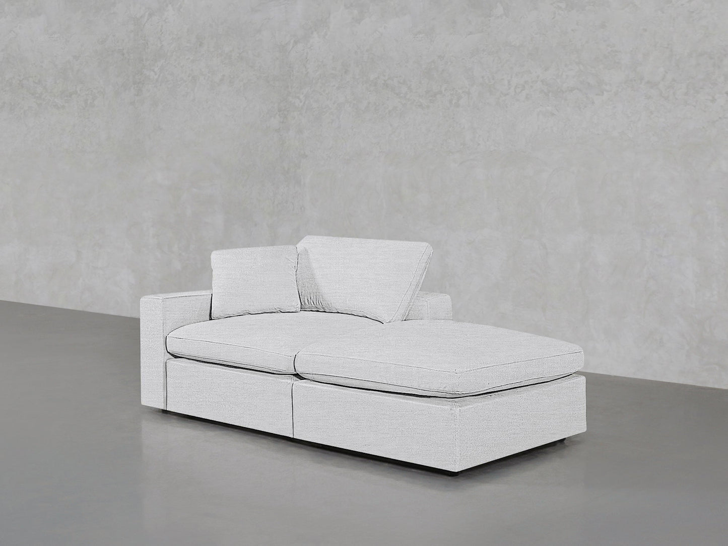 The 2-Seat Modular Lounger Loveseat, in modern light gray upholstery with two seat and back cushions plus an angled cushion, sits on a polished gray floor against a textured gray wall.