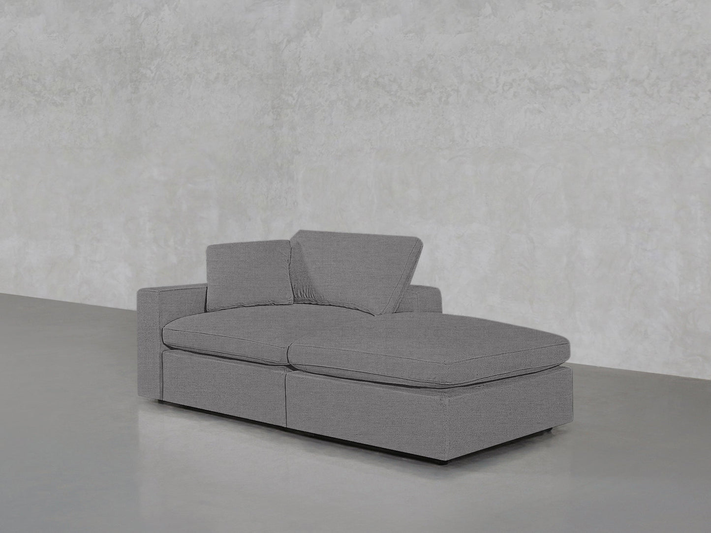 The 2-Seat Modular Lounger Loveseat, in modern gray with two seat and back cushions plus an extended chaise, sits on a smooth gray floor against a textured light gray wall.