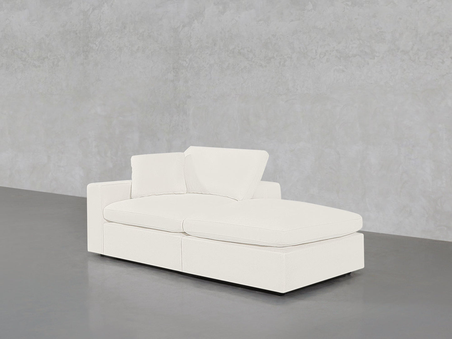 A 2-Seat Modular Lounger Loveseat with two cushions sits in a minimalistic room featuring light gray concrete walls and a smooth gray floor.