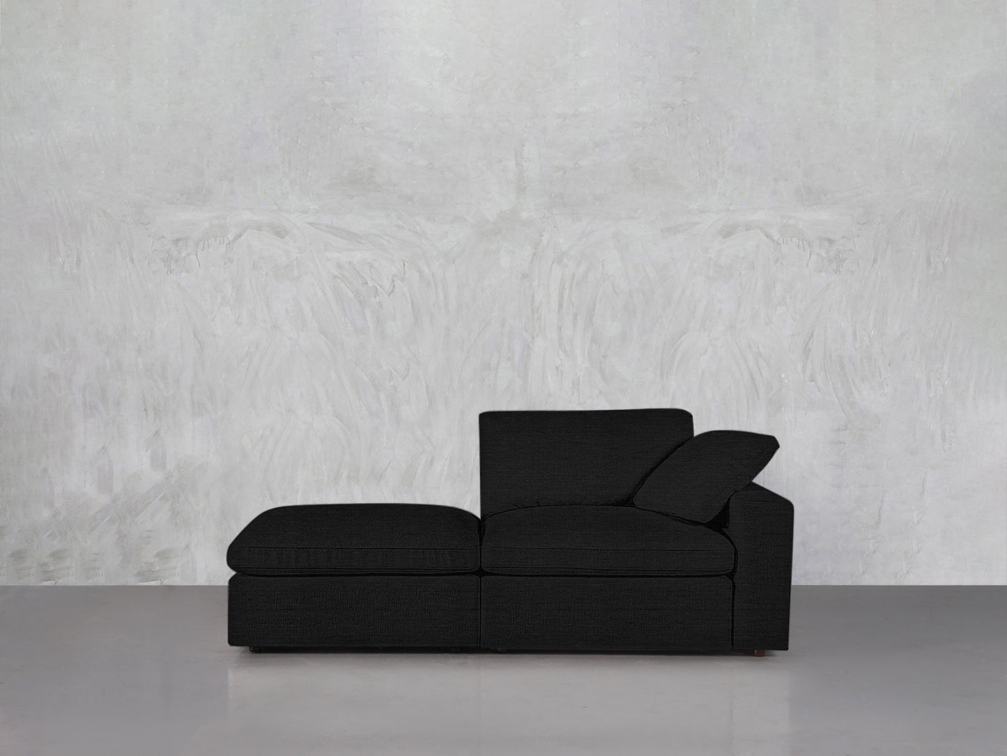 A black 2-Seat Modular Lounger Loveseat with a chaise sits against a light gray textured wall on a glossy gray floor. The minimalist room features only the loveseat, emphasizing its modern design.