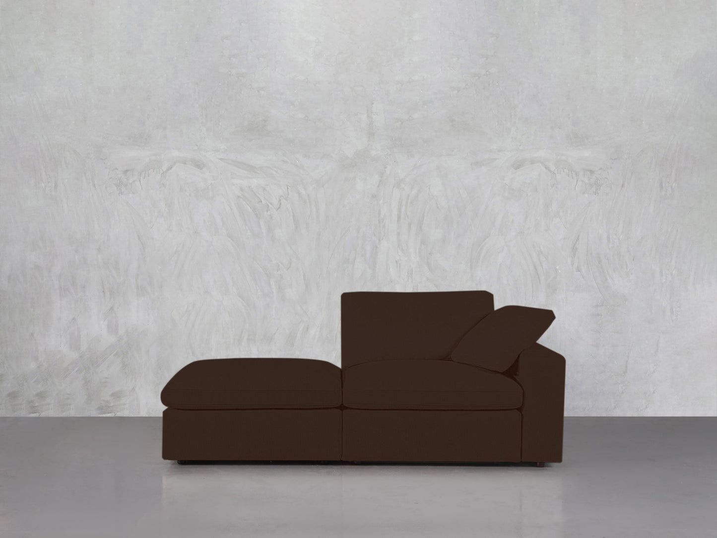The 2-Seat Modular Lounger Loveseat in dark brown stands against a textured light gray wall on a smooth gray floor. One cushion is angled for back support, creating a minimalist and uncluttered setting.