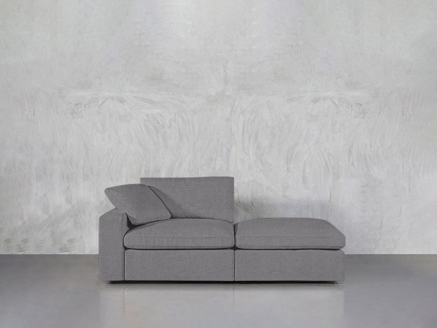A 2-Seat Modular Lounger Loveseat in modern gray with two seat cushions and back pillows is placed against a textured light gray wall on a smooth gray floor.