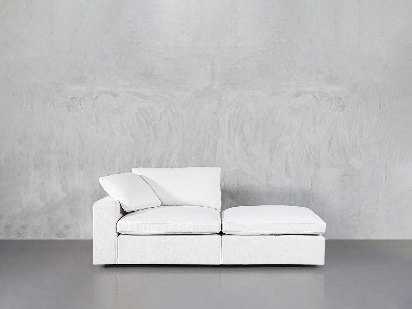 A 2-Seat Modular Lounger Loveseat with two cushions is placed against a light gray textured wall on a smooth gray floor.