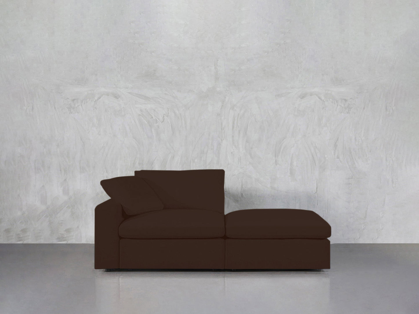 A 2-Seat Modular Lounger Loveseat with a pillow sits against a textured light gray wall and a smooth, reflective gray floor.