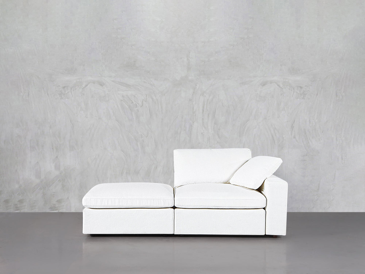 A 2-Seat Modular Lounger Loveseat in modern white with clean lines sits against a light gray textured wall on a smooth gray floor, featuring one armrest and a single back cushion.