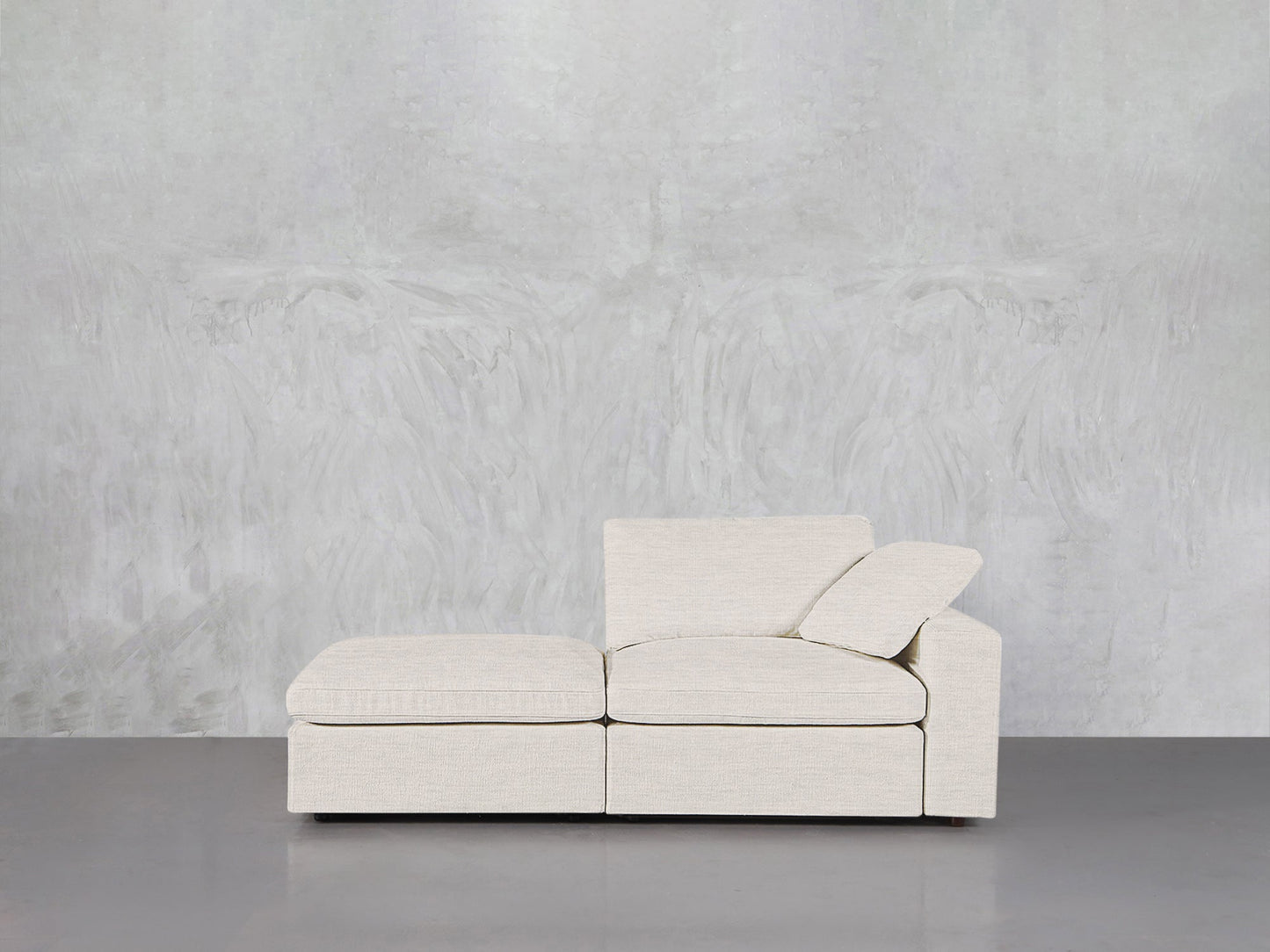 The 2-Seat Modular Lounger Loveseat in light beige, featuring an ottoman and angled backrest, sits before a textured light gray wall on a smooth gray floor.
