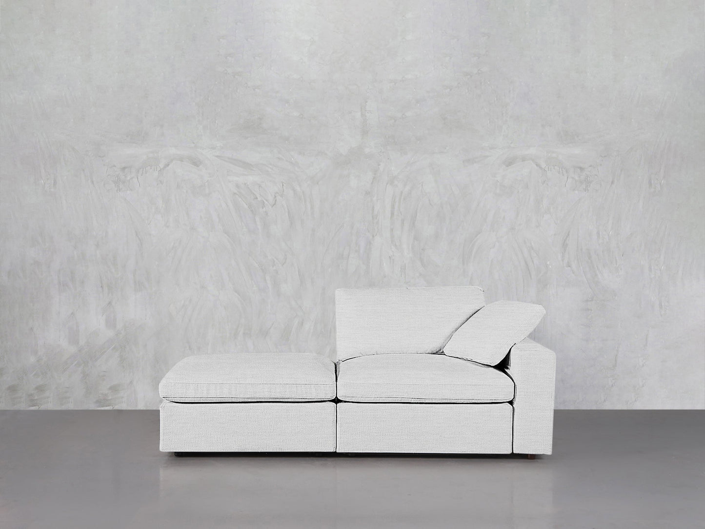 A modern 2-Seat Modular Lounger Loveseat in light gray with one armrest and loose cushions sits against a textured, light gray wall on a smooth, gray floor.