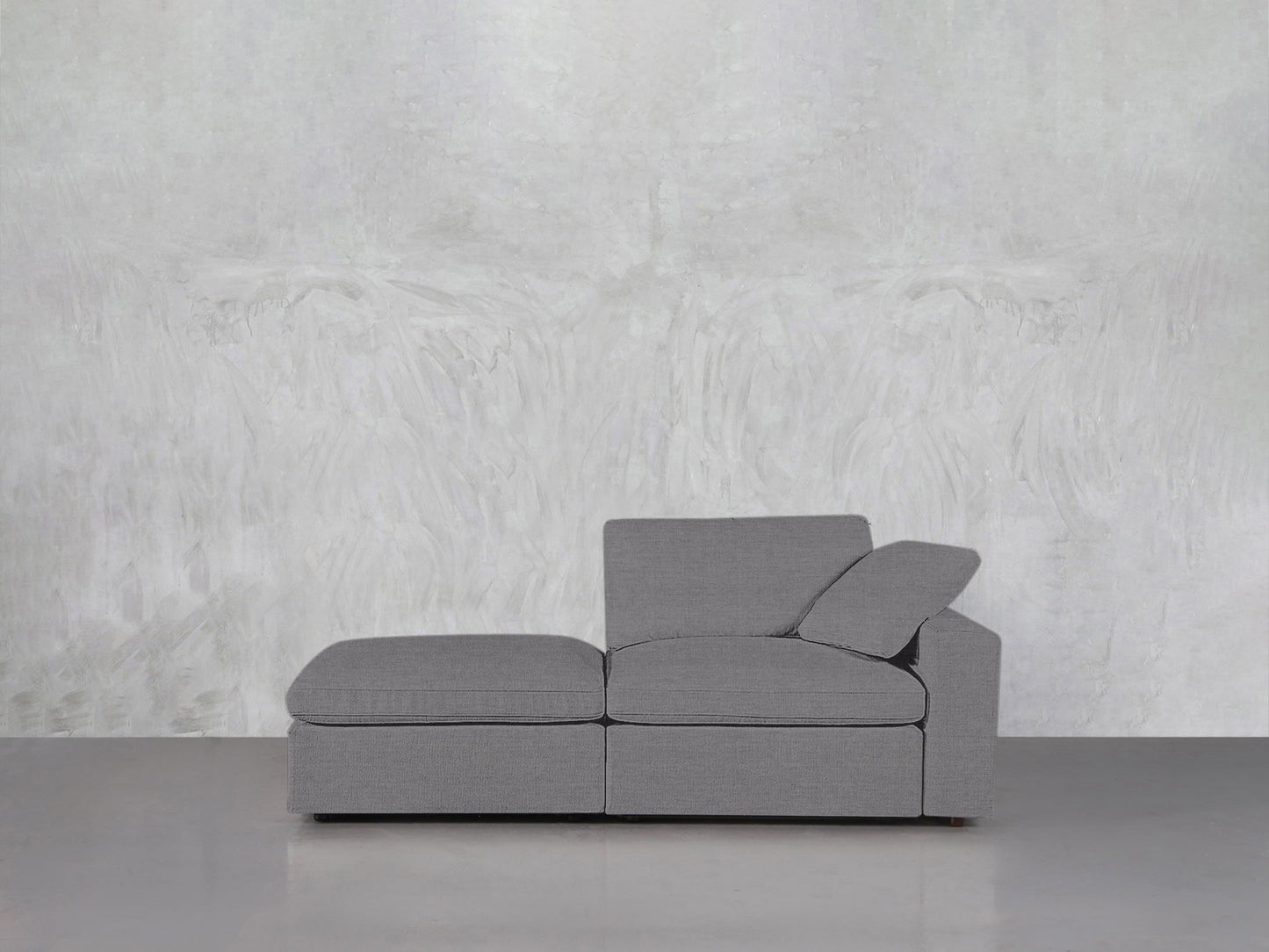 The 2-Seat Modular Lounger Loveseat, featuring clean lines and an angled cushion, is set on a smooth light gray floor against a textured white and gray abstract wall.