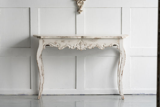 The Slimline wash Console Table, featuring a white washed finish, stands against a white paneled wall, adding shabby chic or French country style to your hallway, living room, or bedroom.