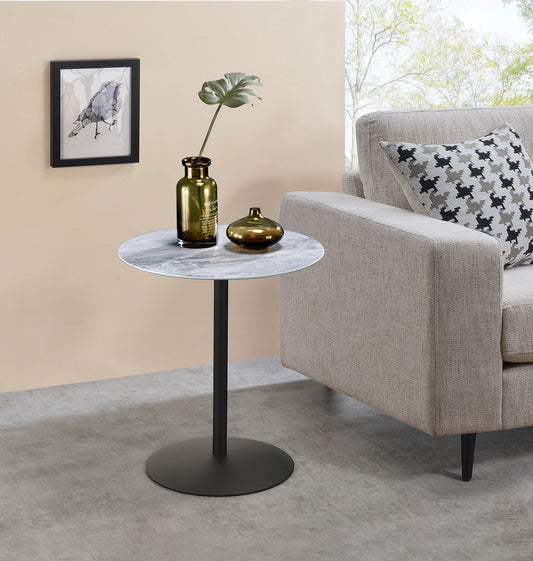 The Circa 17.5" End Table with marble-textured top and a black metal base stands beside a beige sofa. Two vases, one holding a leafy branch, sit on the table beneath a framed bird artwork on the beige wall above.