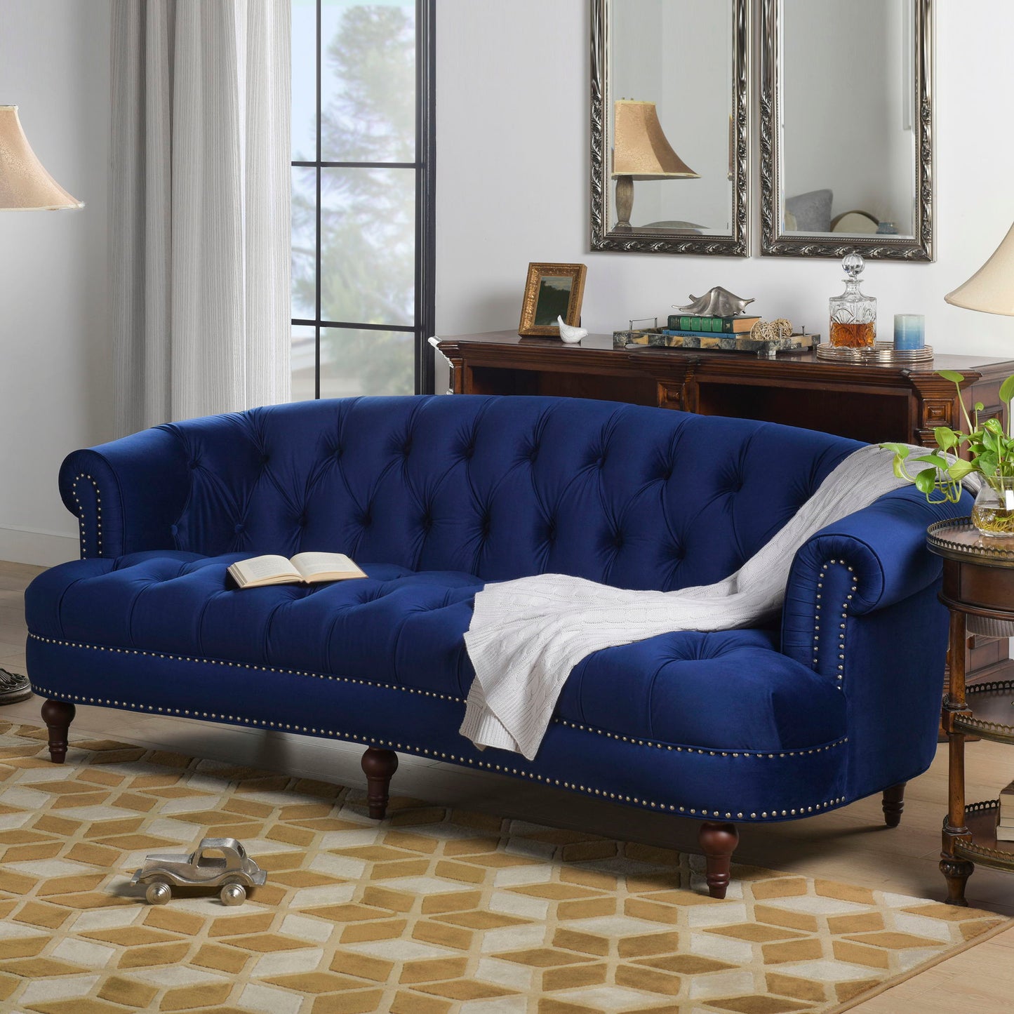 The La Rosa - Victorian Chesterfield Tufted Sofa in blue velvet, featuring studded trim, is styled with a white throw and open book. The cozy living room includes a geometric rug, wooden furniture, lamps, mirrors, and a window.