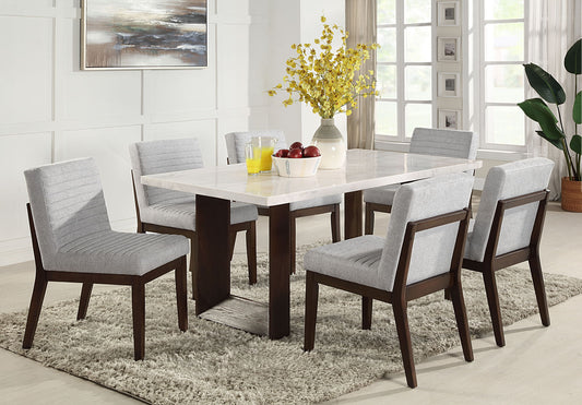 A modern dining room features the Edwyn Marble Top Dining Table - Natural, six gray upholstered chairs, a beige rug, and a yellow flower centerpiece. A bowl of apples and a jug of orange juice sit on the table below large sunlit windows.
