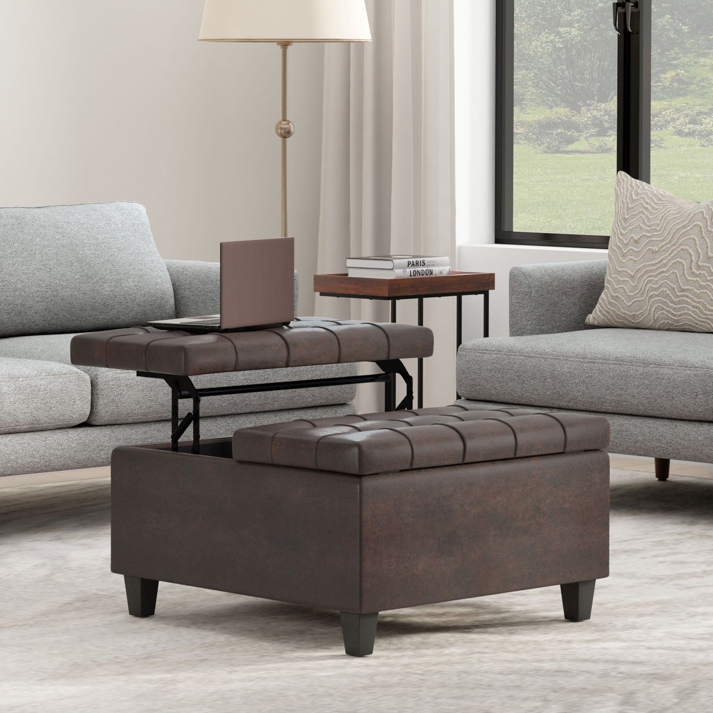 The Harrison Upholstered Coffee Table Storage Ottoman with a lift-top tray sits on a light rug between two gray sofas, with a lamp, books, and a window framed by white curtains in the background.