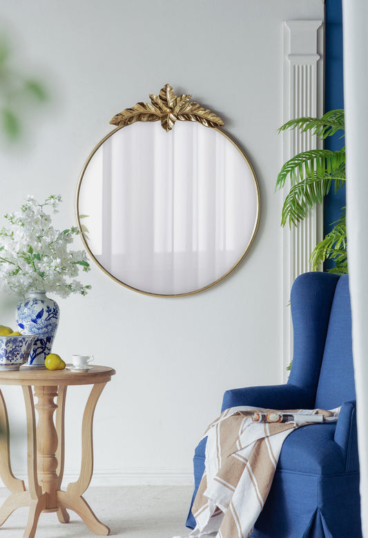 A cozy living room corner with a blue armchair, a Large Round Wall Mirror With Metal Frame in gold, a light wood side table topped with a vase of white flowers, and a potted plant set against a white wall.
