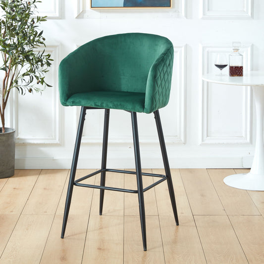 A Counter Height Bar Stool, Metal Legs, Velvet Upholstered Set in Black / Green stands on a light wood floor beside a small white table with a wine glass and decanter, with a leafy potted plant in the background.