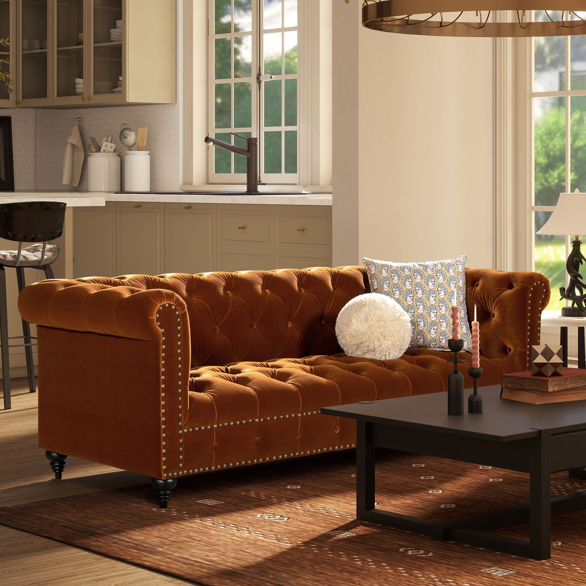 A cozy living room features the Alto Tufted Chesterfield Sofa in rust, two decorative pillows, a black coffee table with candles, a patterned rug, and beige kitchen cabinets in the background. Sunlight streams through tall windows.