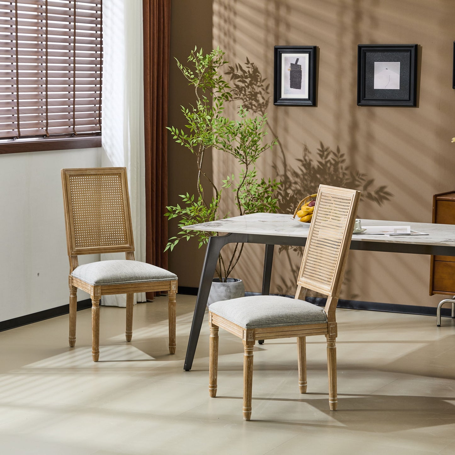 Two French Country Dining Chairs with rectangular cane backrests and smooth upholstered seats are placed by a modern table in a sunlit room, enhanced by a potted plant, wall art, and wooden blinds for a cozy minimalist touch.