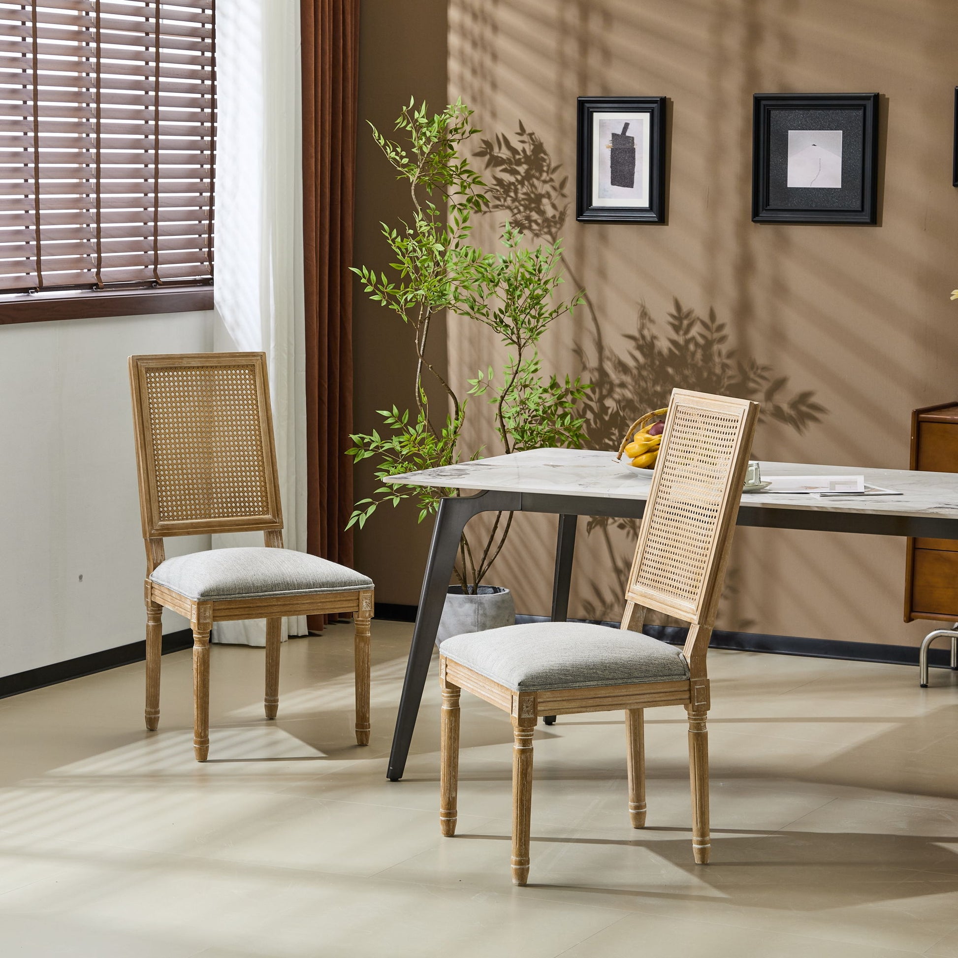 Two French Country Dining Chairs with rectangular cane backrests and smooth upholstered seats are placed by a modern table in a sunlit room, enhanced by a potted plant, wall art, and wooden blinds for a cozy minimalist touch.