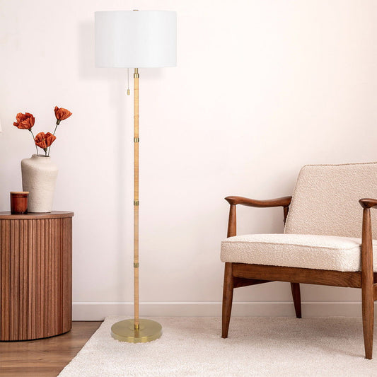 A modern living room corner with a beige armchair, the Stellar Linen Shade Floor Lamp in gold, natural wood, rattan, and white, a round wooden side table topped with a vase of orange flowers on a light-colored rug.