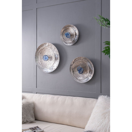 The Textured Oversized Disc Wall Decor For Living Room (Set of 3) – Multicolor is displayed on a gray paneled wall above a beige couch with textured pillows, with a green plant partly visible to the right.