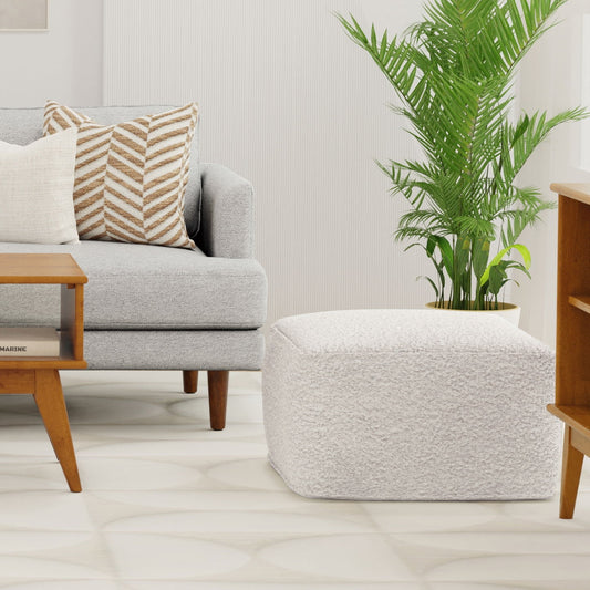 A modern living room with a light gray sofa, patterned and solid pillows, a wooden coffee table, the Chantal Square Pouf in Ivory, a large green potted plant, and a neutral rug on the floor.