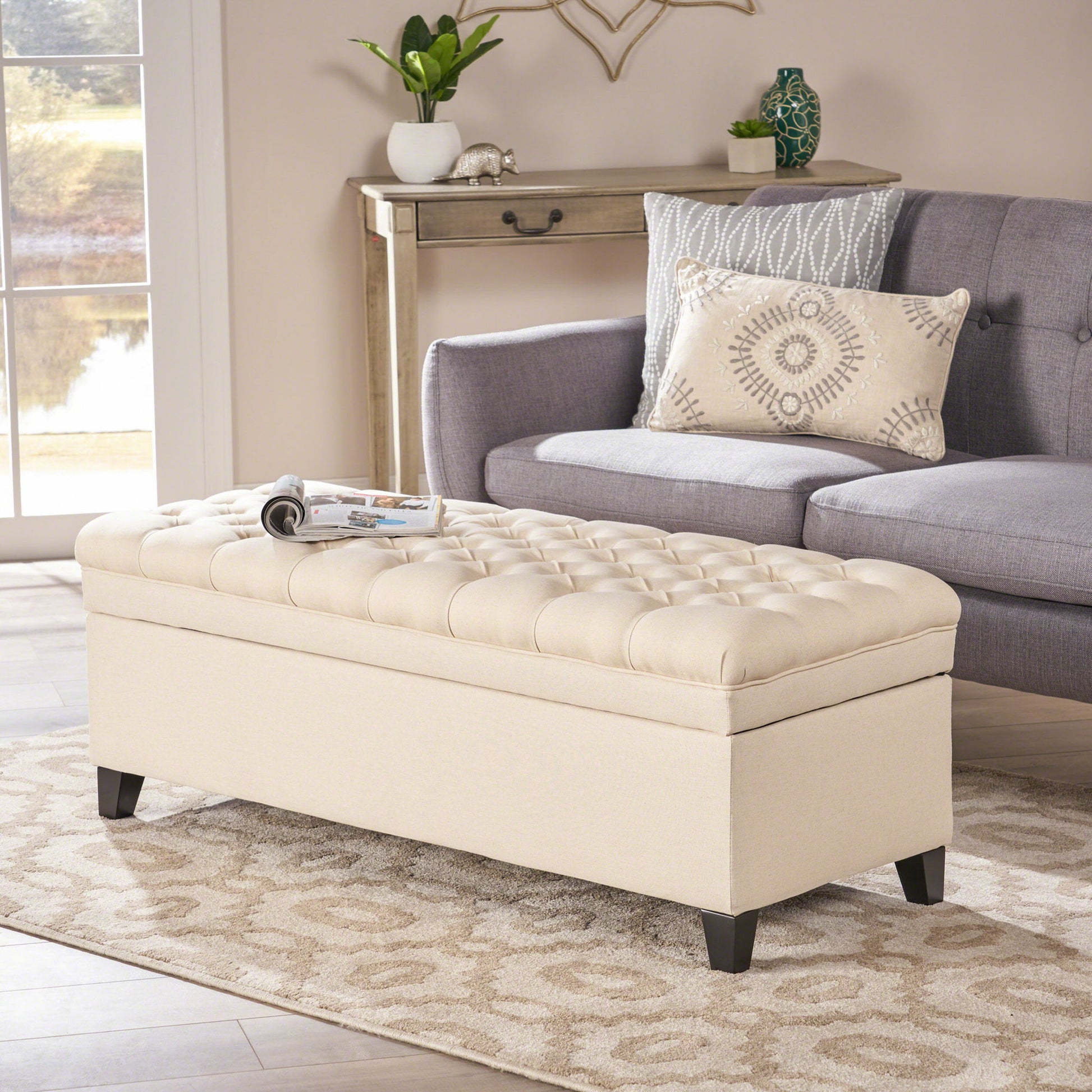 The Juliana Storage Ottoman with Birch Legs in beige stands on a patterned rug in front of a gray sofa with patterned cushions, near a window and a console table with decor, creating a bright and cozy living room.