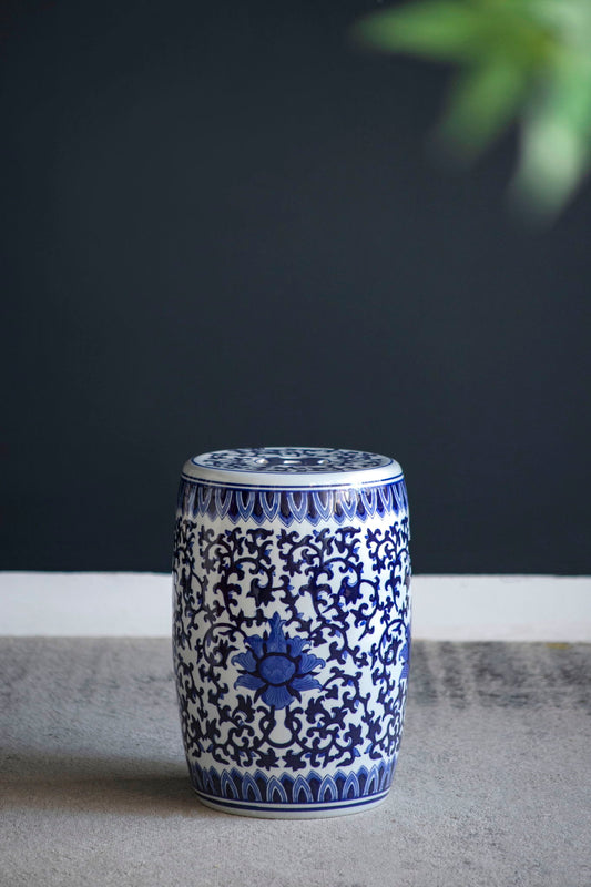 The Ceramic Garden Stool, Small Side Table in white and blue with intricate floral patterns sits on a gray floor before a dark wall, while a green plant appears partially in the top right corner.