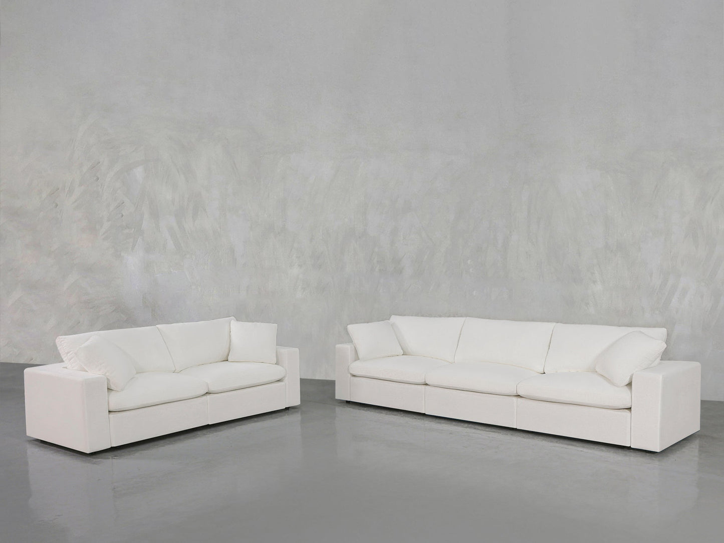 The 3-2 Sofa Set, featuring modern white sofas with plush cushions, is arranged on a shiny gray floor against a textured light gray wall in a minimalistic room.
