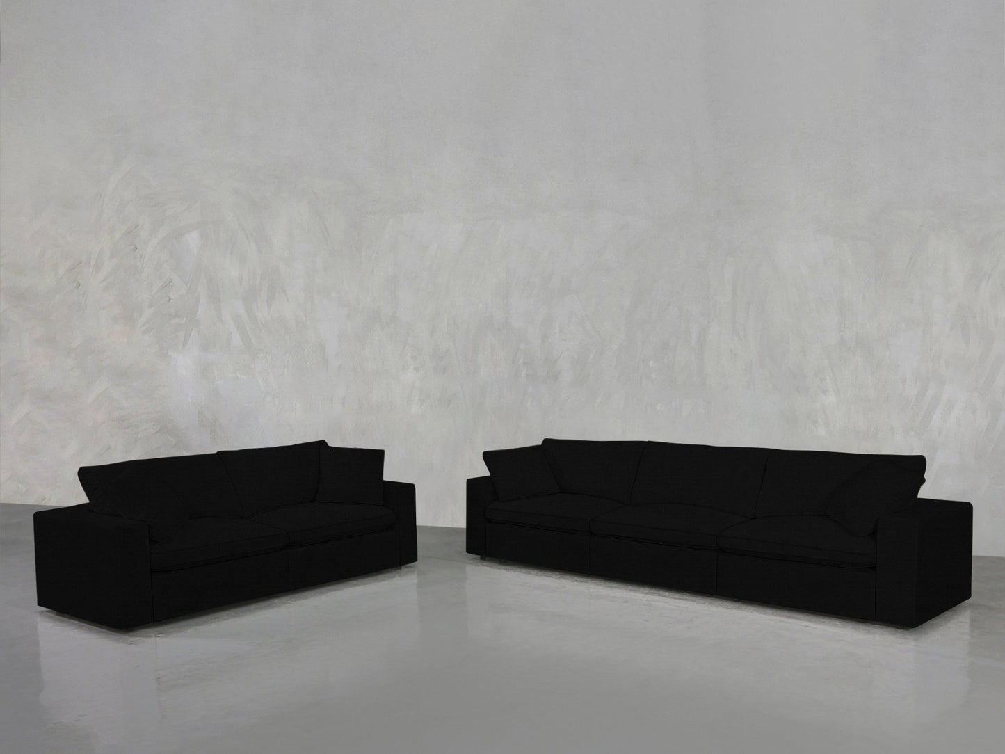 The 3-2 Sofa Set, featuring one larger and one smaller black sofa, sits on a shiny gray floor against a light gray textured wall in a minimal, empty room.