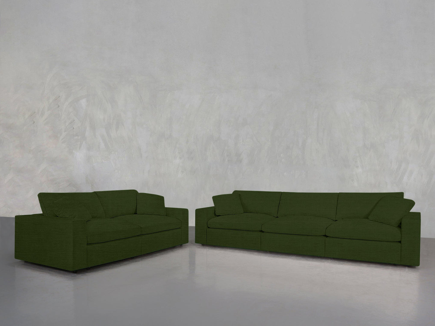 The 3-2 Sofa Set, featuring larger and smaller dark green sofas, sits on a smooth grey floor against a textured light grey wall in a spacious, minimalistic room.