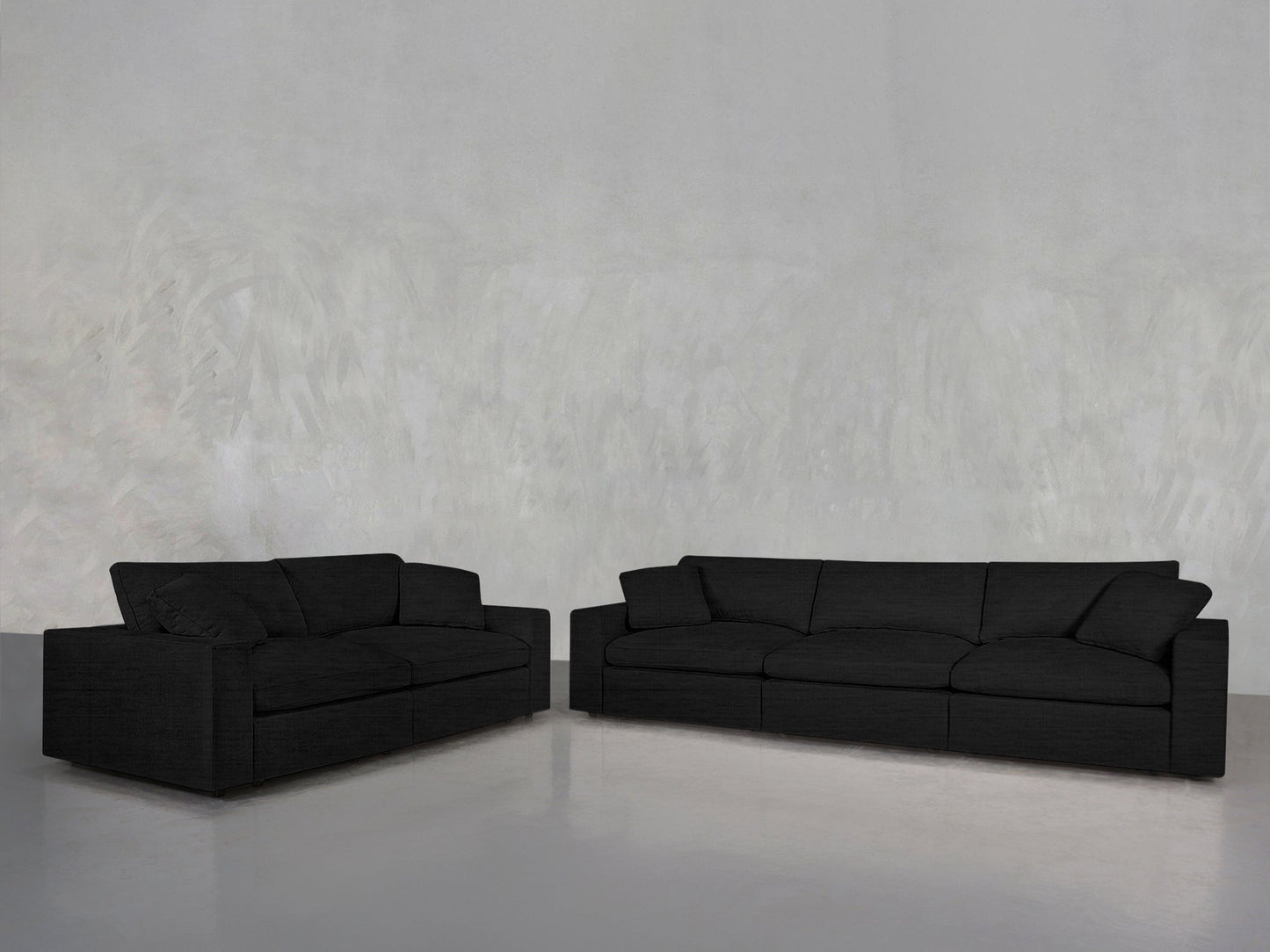 The 3-2 Sofa Set, featuring modern black fabric two- and three-seater sofas, is arranged in an empty room with a light gray textured wall and a smooth gray floor.