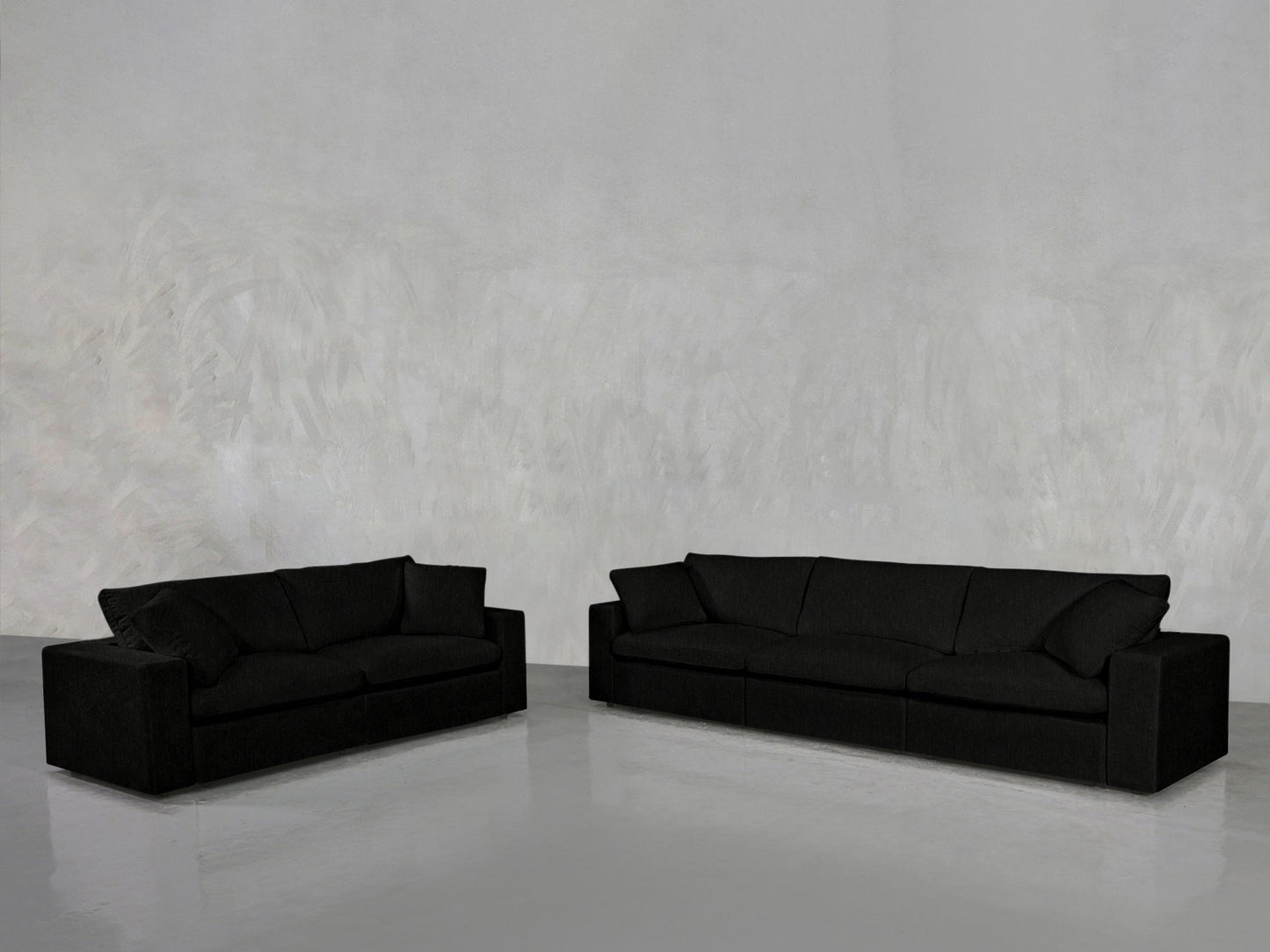 The 3-2 Sofa Set, in sleek black, stands against a light gray textured wall on a polished floor. The minimalistic, empty room highlights the contemporary design of the sofas.