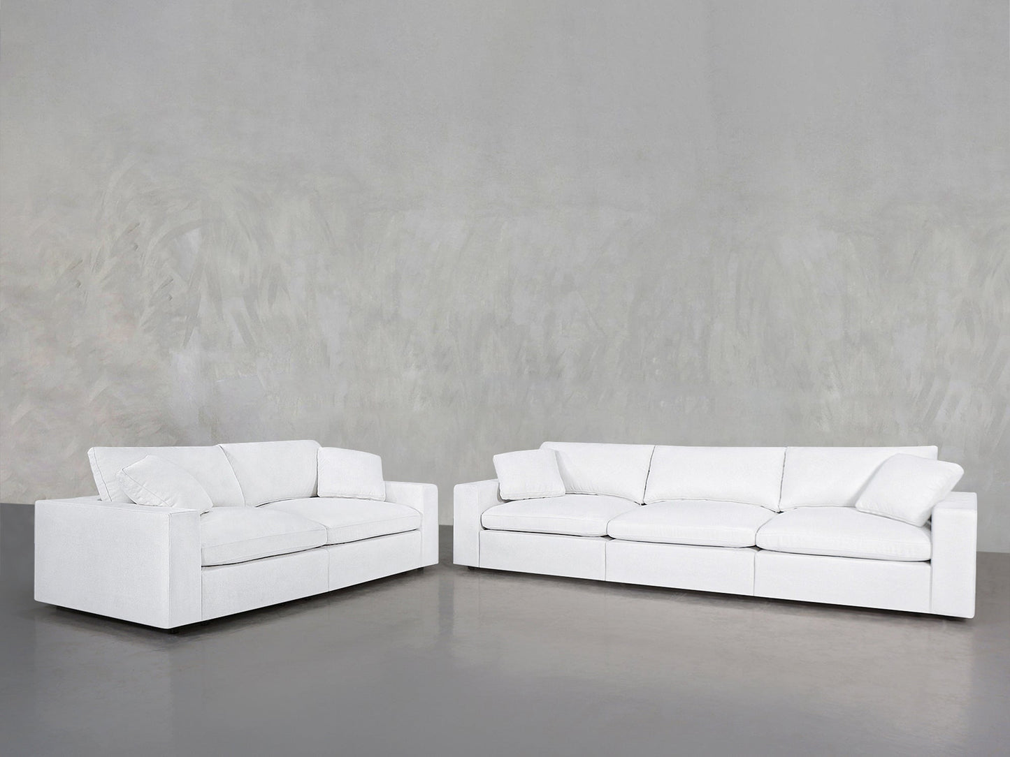 The 3-2 Sofa Set, featuring two sleek white sofas with plush cushions, is arranged in an L-shape on a smooth grey floor and set against a textured light grey wall.