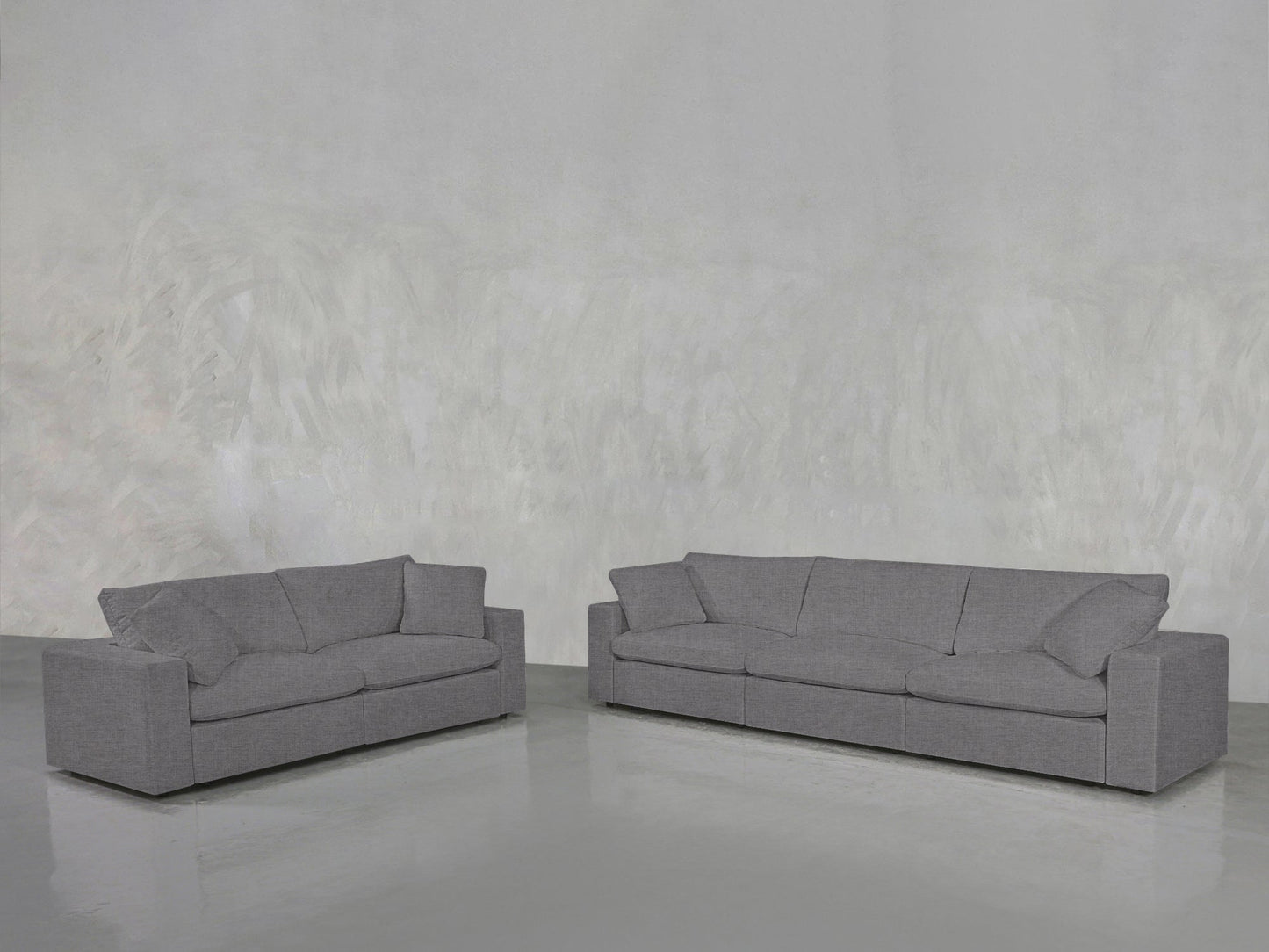 The 3-2 Sofa Set features a modern two-seater and three-seater in gray with clean lines and plush cushions, placed on a polished floor against a textured light gray wall for a minimalistic look.