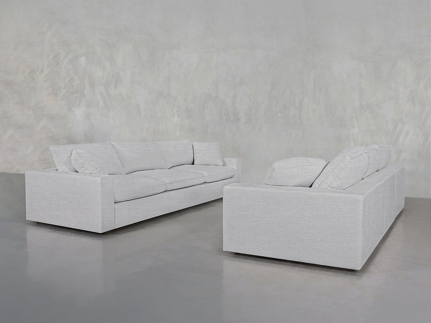 Two contemporary 3-3 Sofa Set pieces in light gray, featuring boxy shapes and plush cushions, are arranged opposite each other in a minimalist room with smooth gray concrete flooring and a textured gray wall.