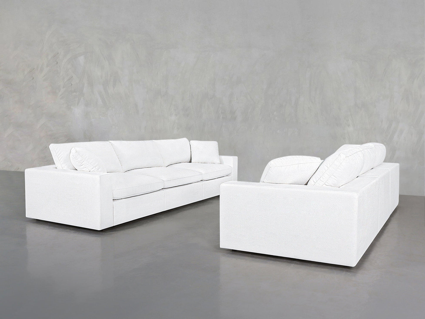 Two 3-3 Sofa Set pieces with modern white upholstery, clean lines, and large cushions sit on a smooth gray floor against a textured light gray wall, placed at an angle facing each other.