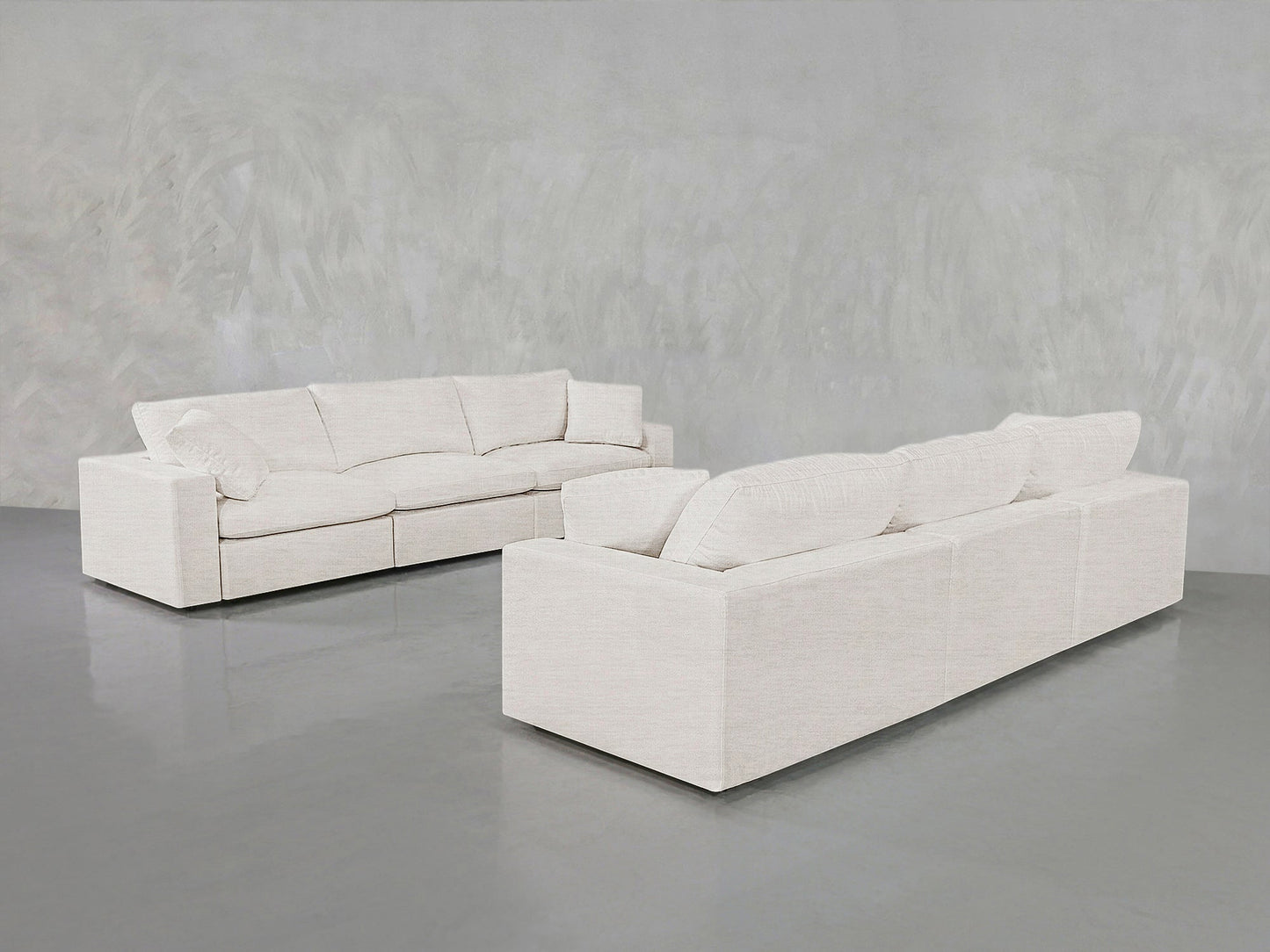 Two modern 3-3 Sofa Sets in light beige are arranged in an empty room with a smooth gray floor and textured light gray walls. These sofas feature plush cushions and a minimalist design.