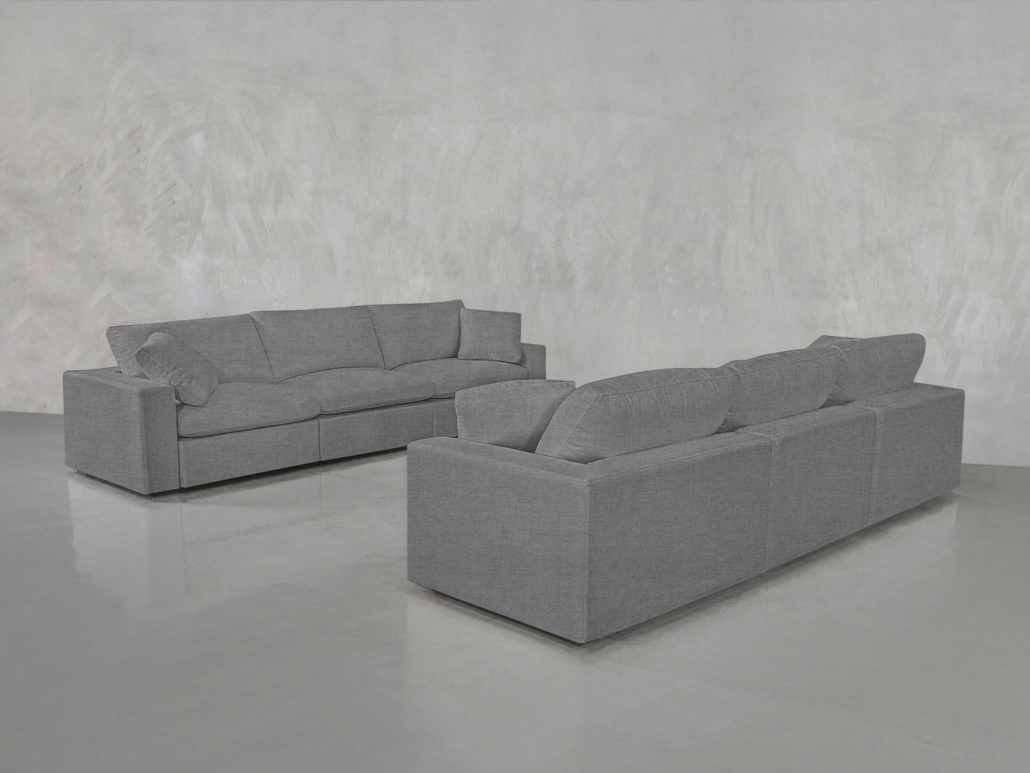The 3-3 Sofa Set, featuring clean lines and plush cushions in modern gray fabric, is arranged in an empty room with a light gray textured wall and polished concrete floor.