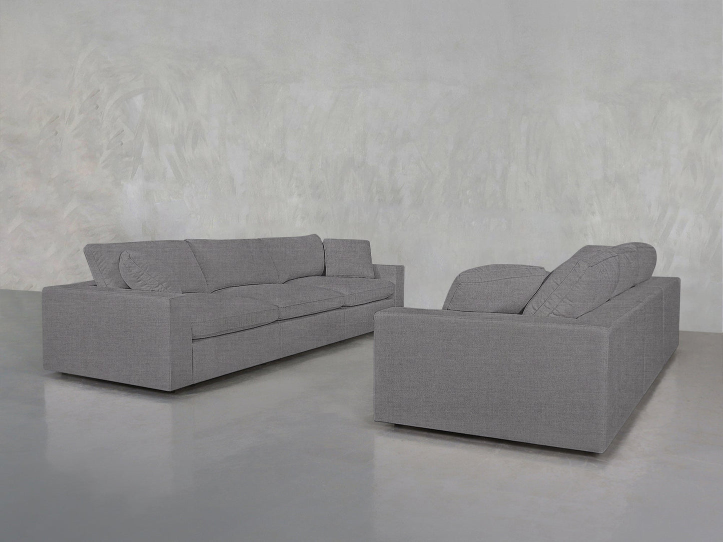 Two 3-3 Sofa Set pieces in modern grey, featuring clean lines and plush cushions, are arranged in an empty room with a light grey textured wall and a polished concrete floor.