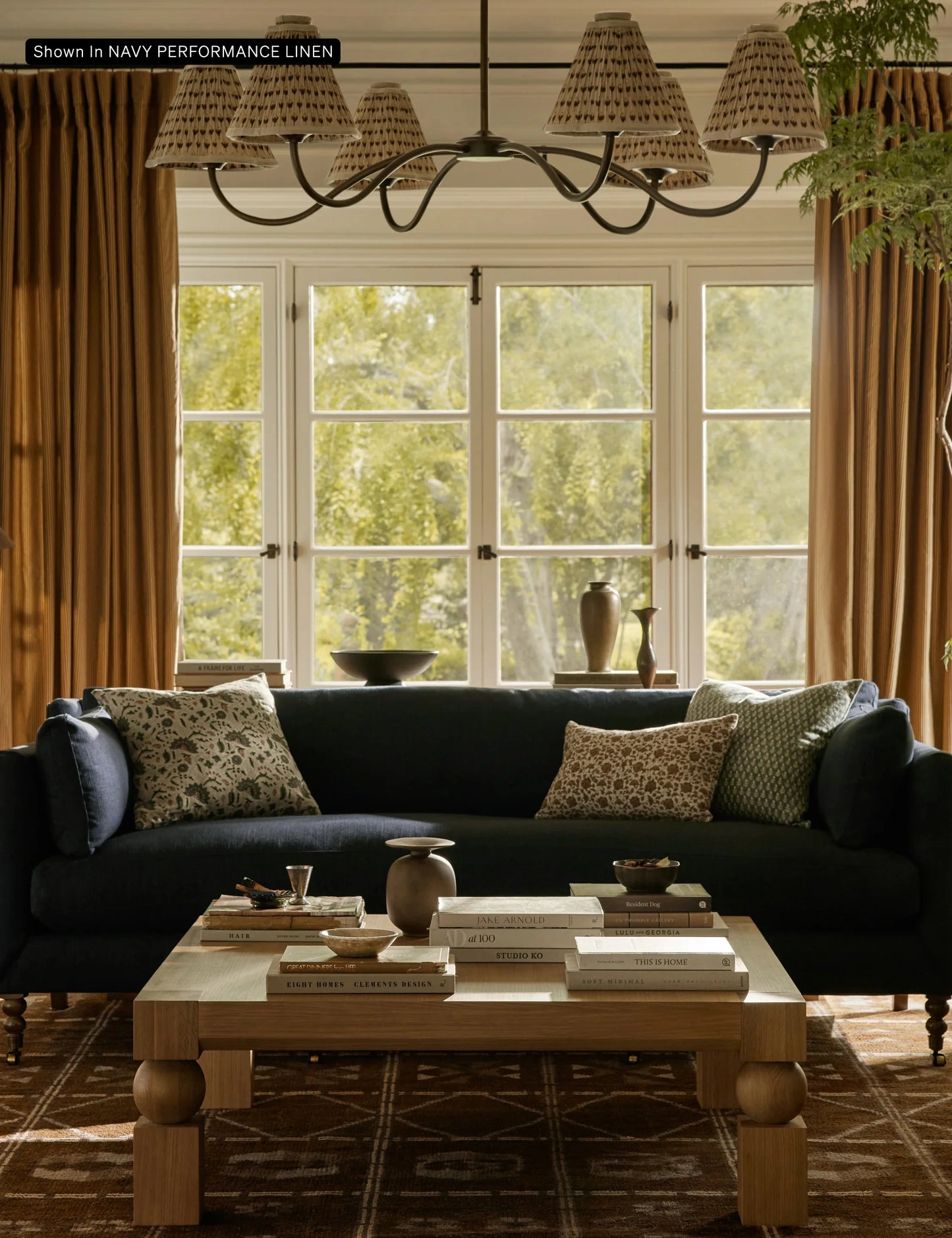 A cozy living room featuring the Fabienne Sofa in dark blue, adorned with patterned cushions. In front of it sits a wooden coffee table holding books and decorative items. Large windows with brown curtains showcase a view of the greenery outside, while a chandelier hangs overhead.