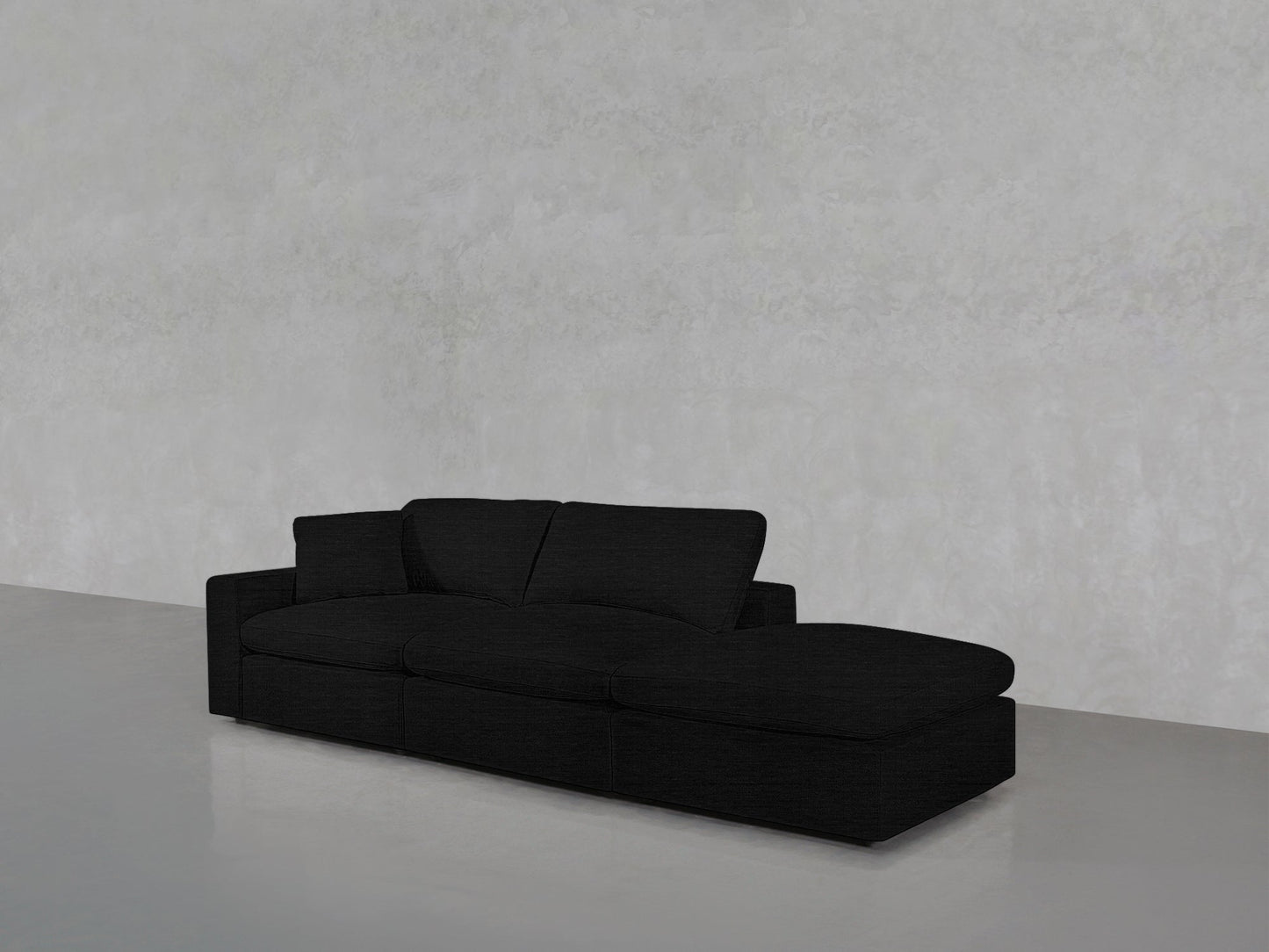 The 3-Seat Modular Lounger Sofa in black, featuring three seat cushions and a chaise lounge, is positioned against a light gray textured wall on a smooth gray floor.