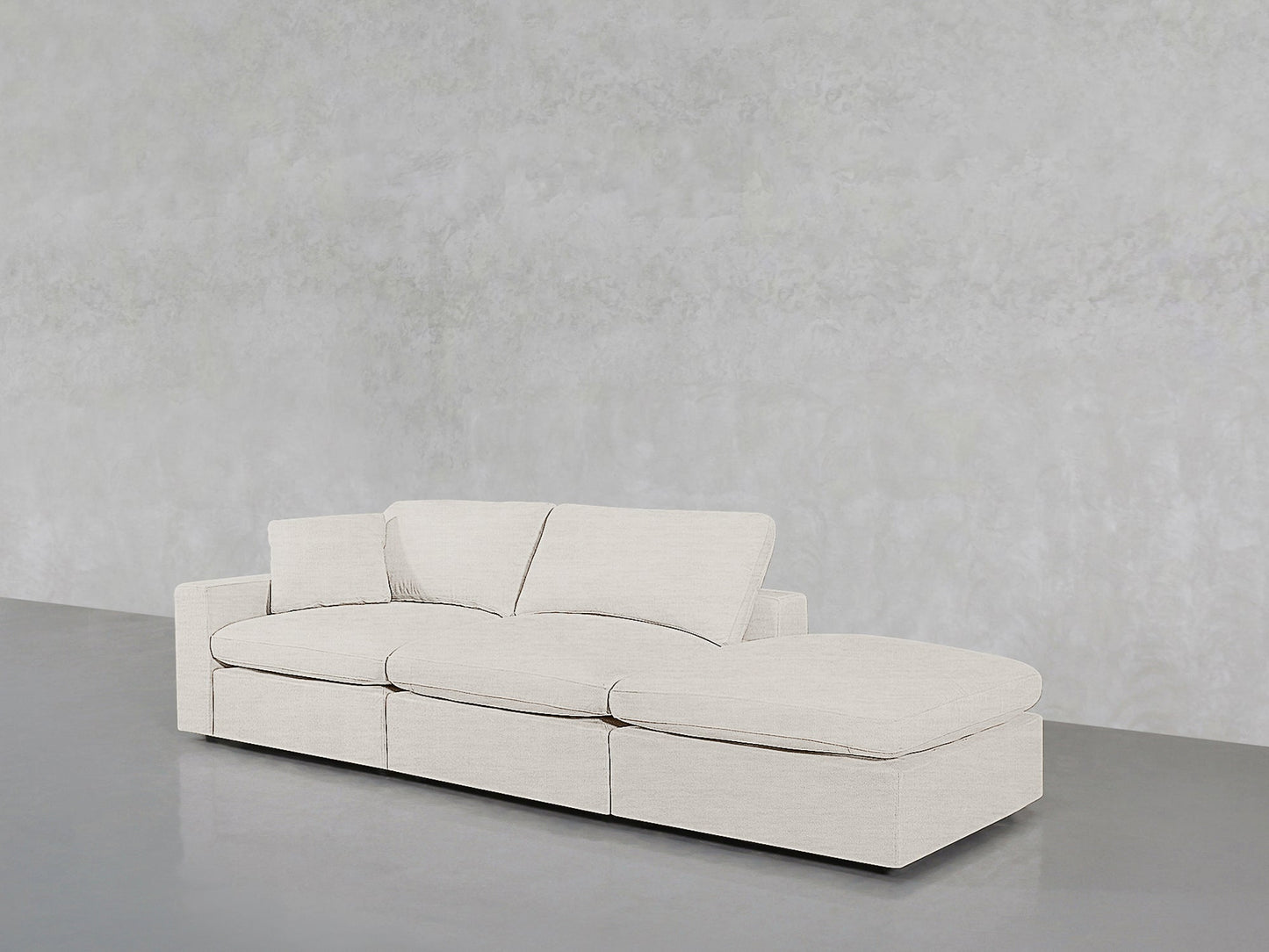 The 3-Seat Modular Lounger Sofa features a modern light beige design with three seat cushions, a right-side chaise, and low square armrests, set against a plain light gray textured wall and gray flooring.