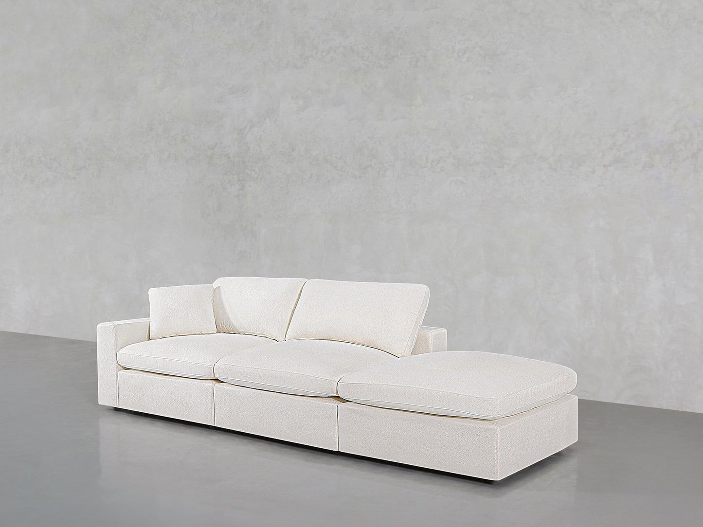 The 3-Seat Modular Lounger Sofa features white upholstery, three seat cushions, and a right-side chaise lounge, set against a light gray textured wall and smooth gray floor.