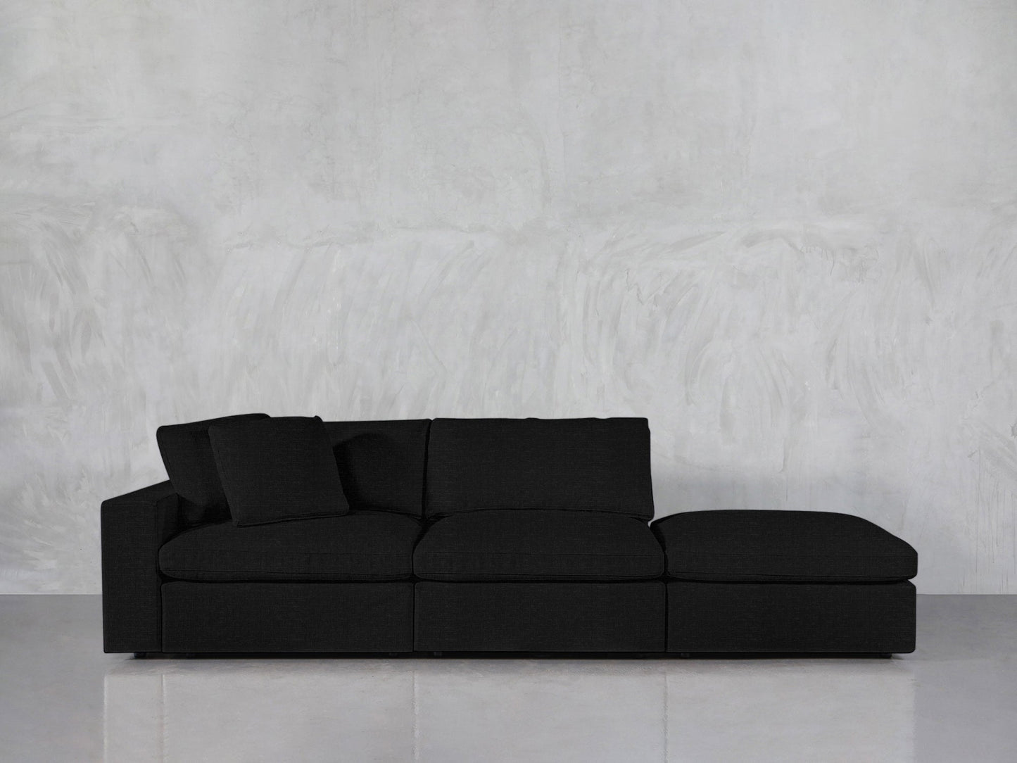 The 3-Seat Modular Lounger Sofa, a modern black sectional with three seat cushions and two black throw pillows, is placed against a light gray textured wall on a smooth concrete floor.
