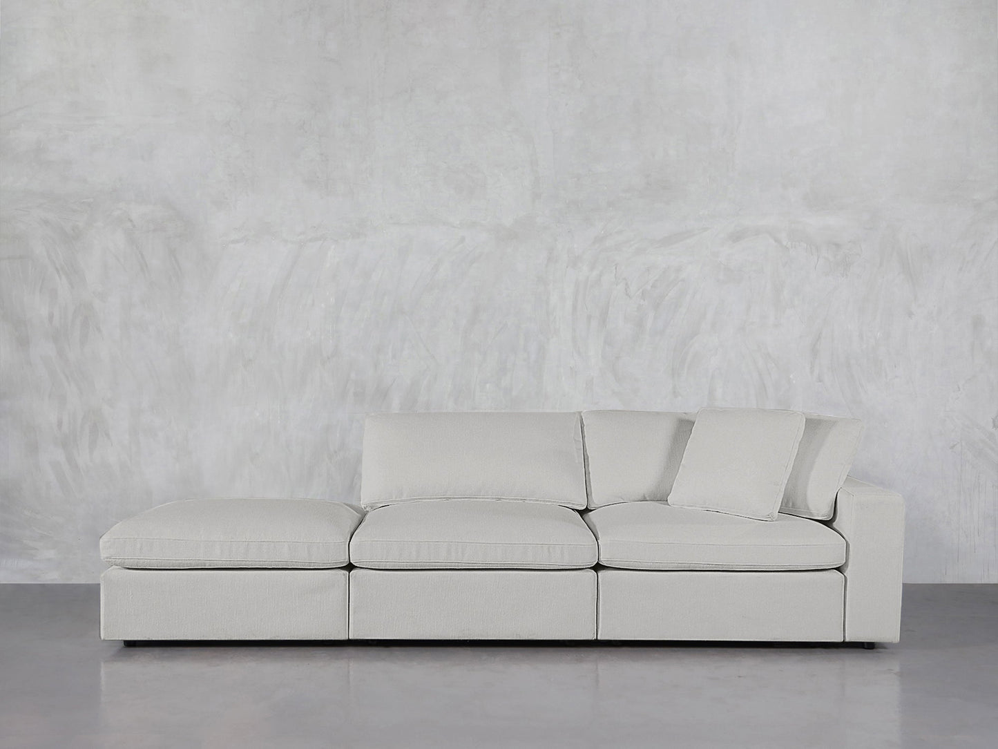 The 3-Seat Modular Lounger Sofa in light gray features clean lines and loose cushions, set against a textured gray wall on a polished floor. One back cushion is tilted against the armrest for a relaxed look.