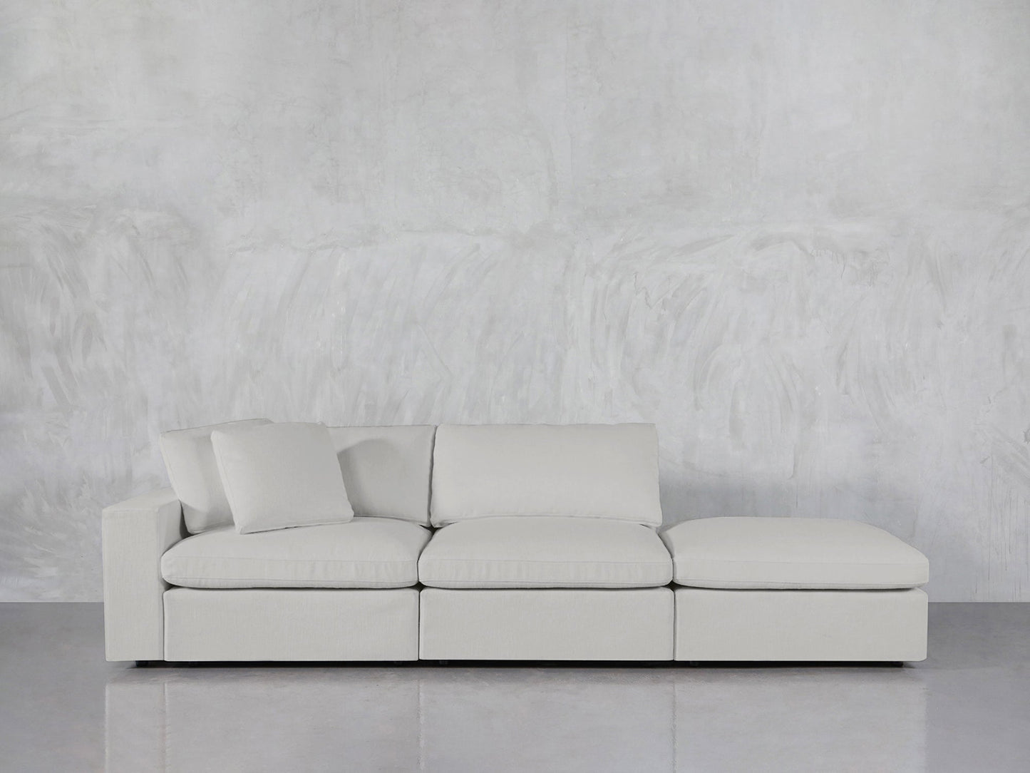 The 3-Seat Modular Lounger Sofa in light gray, with three cushions and a chaise lounge, is set against a textured gray wall on a polished gray floor. A single pillow rests on the left side of the sofa.