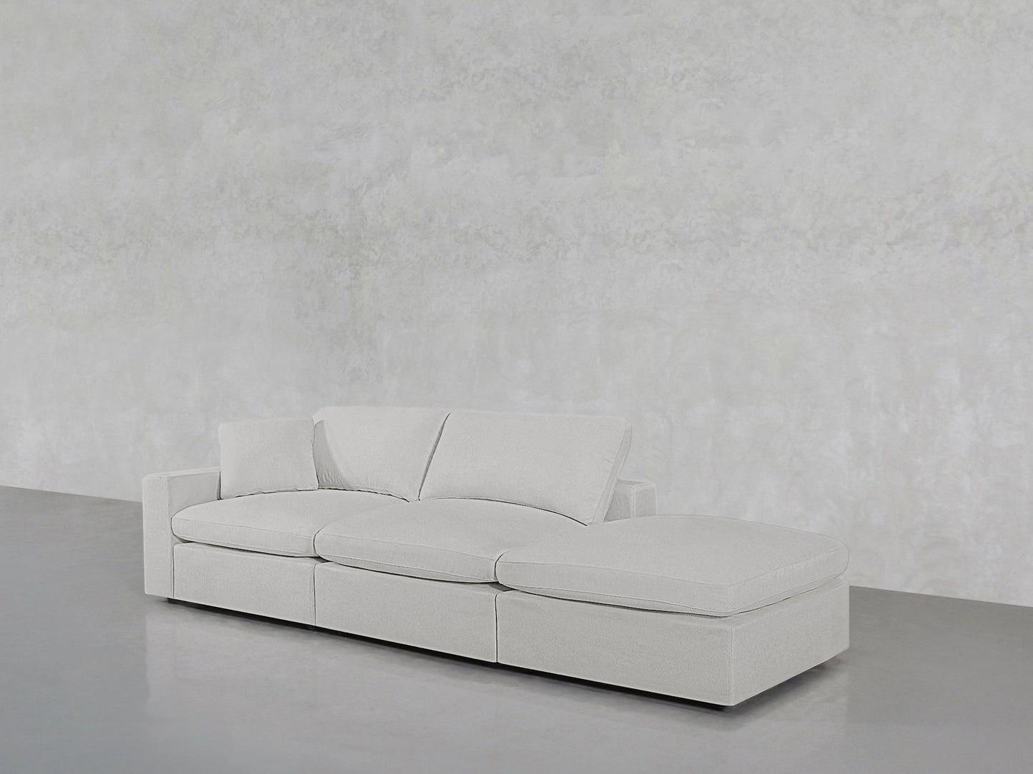 The 3-Seat Modular Lounger Sofa features a modern light gray design with three seat cushions, two back pillows, and a right-side chaise lounge, showcased against a textured light gray wall and smooth gray floor.